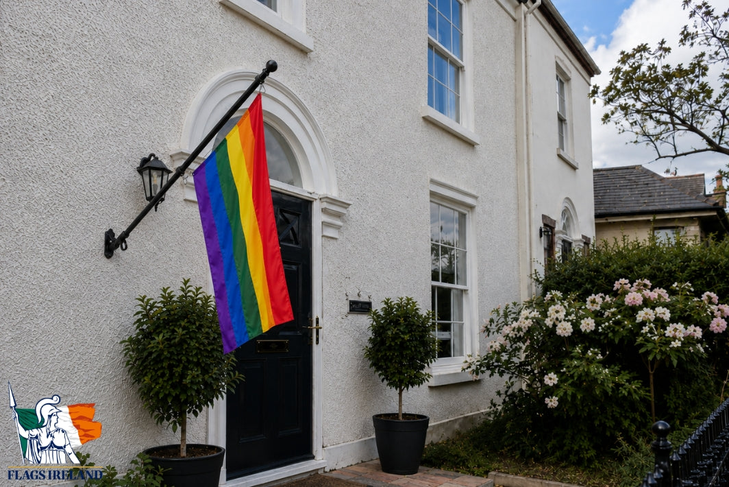 A rainbow pride flag hangs from a black metal house flagpole mounted on the exterior wall of a white stucco house. The scene includes a black front door, potted shrubs, flowering bushes, and part of the neighboring house and sky.