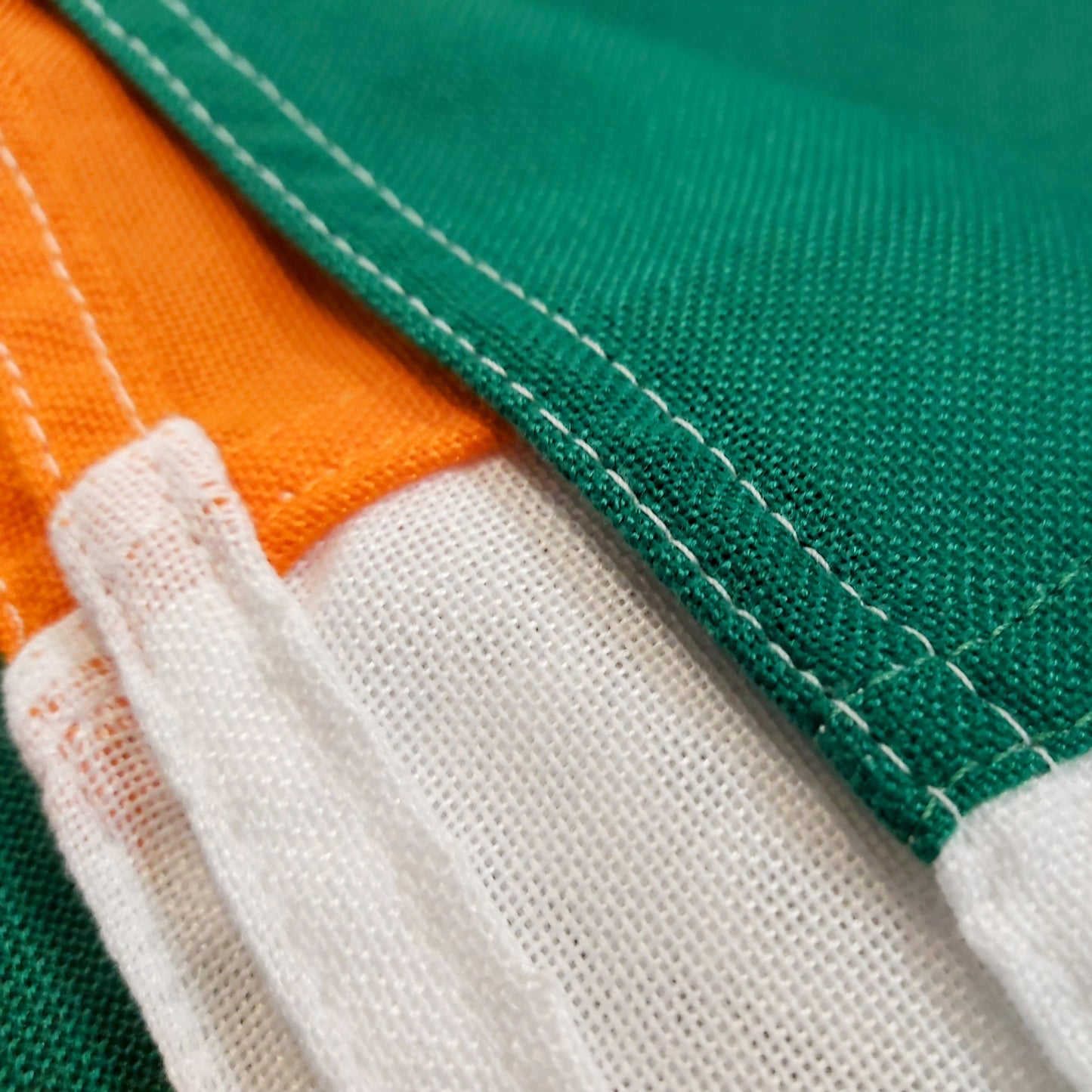 A close-up image of an Irish flag made from heavy woven polyester, showing the high-quality fabric, reinforced stitching, and securely attached hems. The green, white, and orange panels are sewn together with precision.