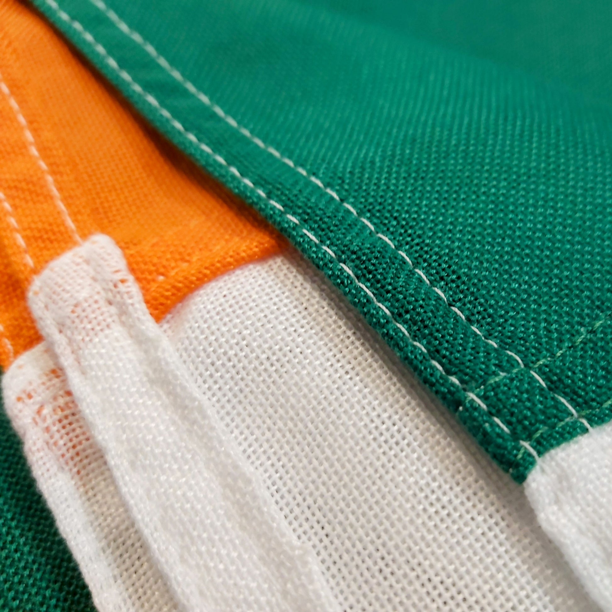A close-up image of an Irish flag made from heavy woven polyester, showing the high-quality fabric, reinforced stitching, and securely attached hems. The green, white, and orange panels are sewn together with precision.