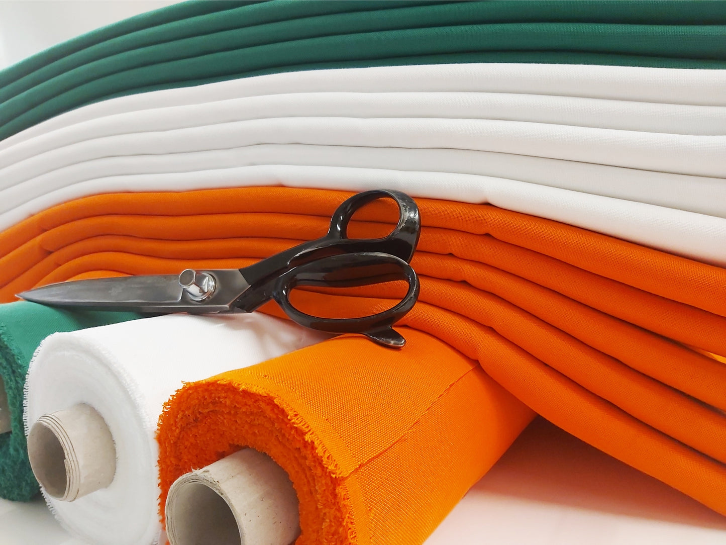 Stacks of green, white, and orange woven fabric rolls used for making Irish tricolour flags. A pair of black tailoring scissors rests on the orange fabric, representing the craftsmanship behind flag production.
