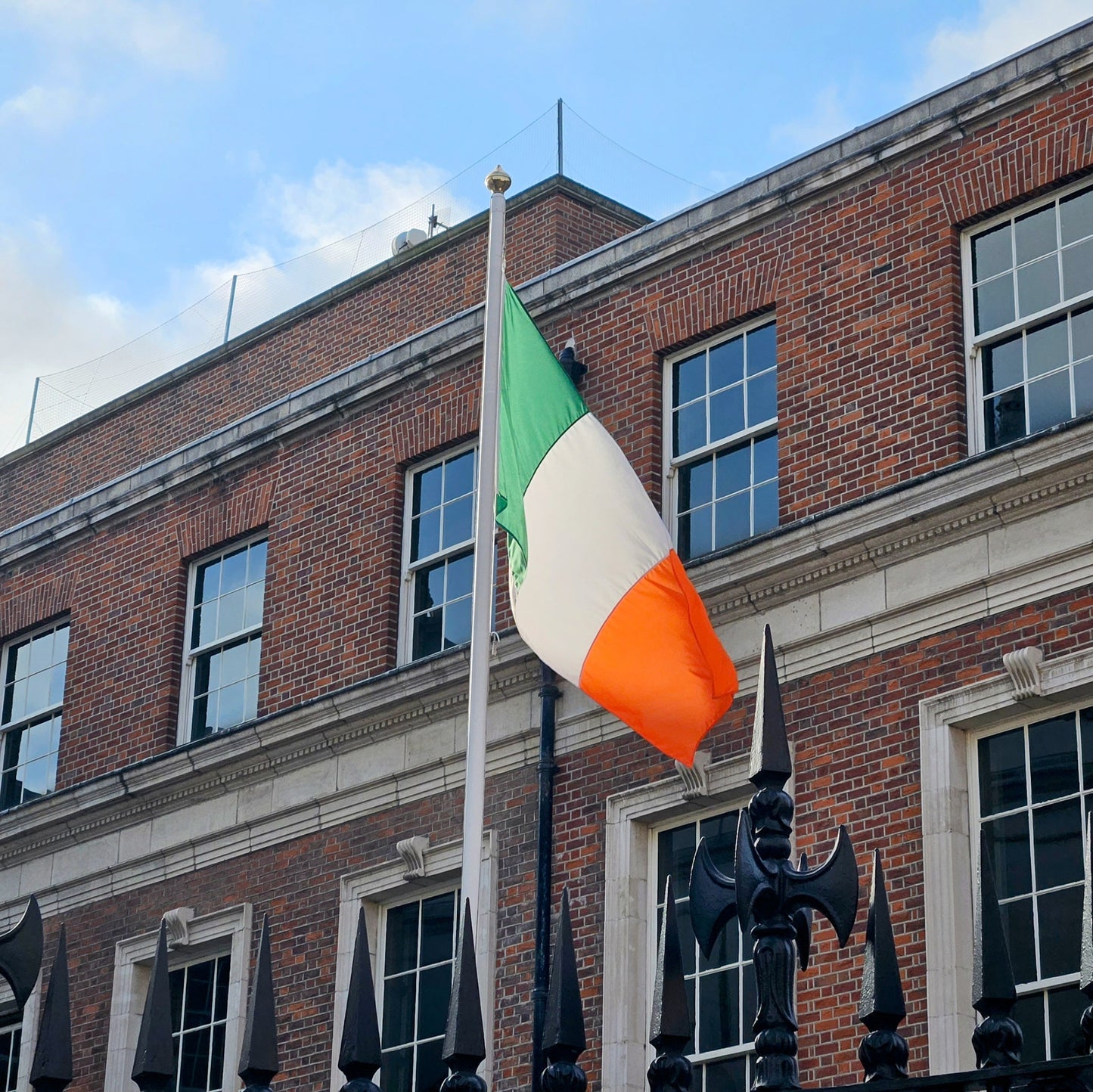 A high-quality Irish tricolour flag flying outside a historic red-brick government building. The flag is mounted on a tall flagpole, waving in the wind against a blue sky. The durable material and stitching are visible, showcasing its premium quality.