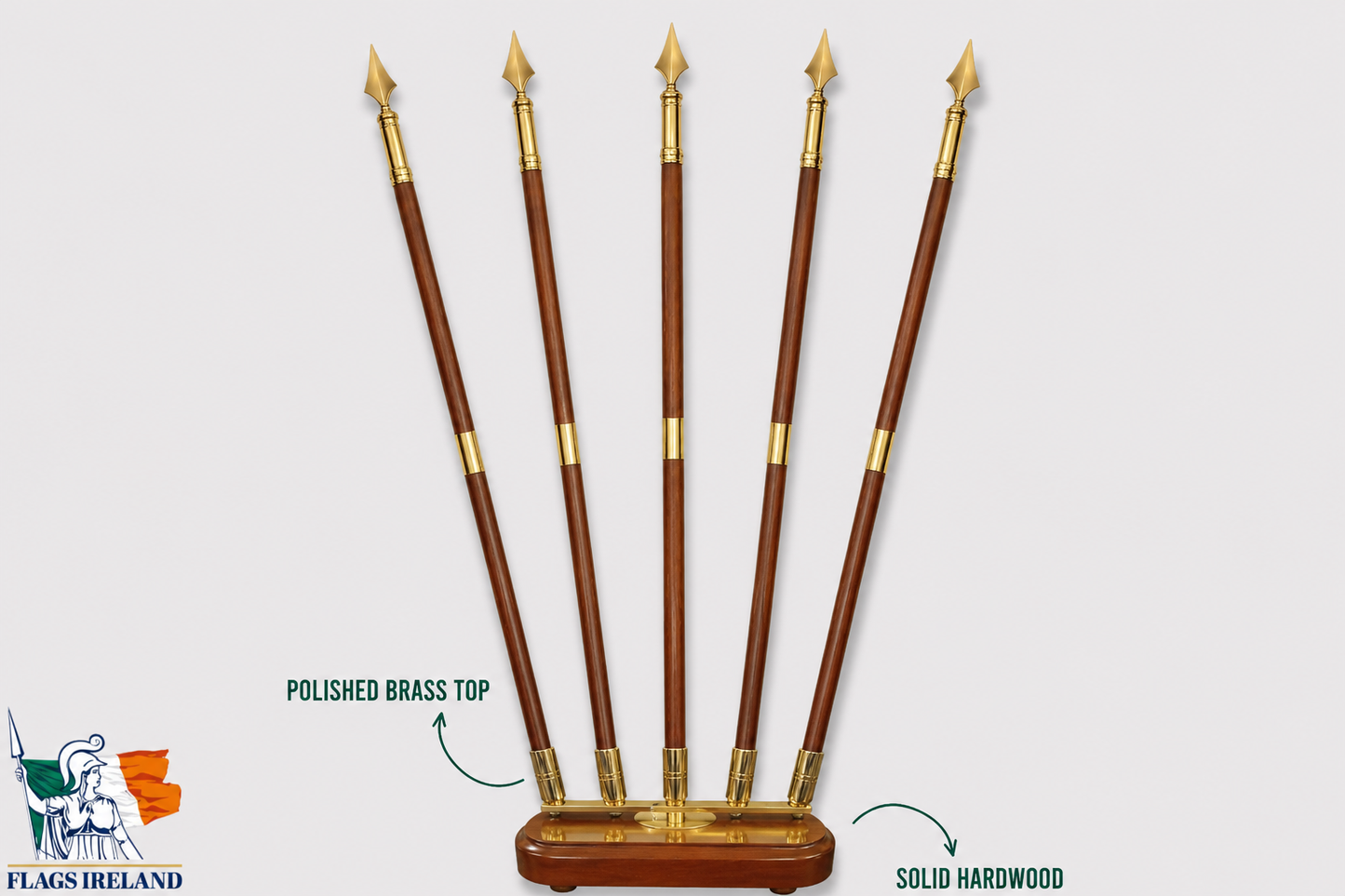 Five polished hardwood flag poles with polished brass tips arranged upright on a wooden base against a light gray background, with text callouts reading 'POLISHED BRASS TOP' and 'SOLID HARDWOOD'. A 'FLAGS IRELAND' logo appears in the lower left corner.