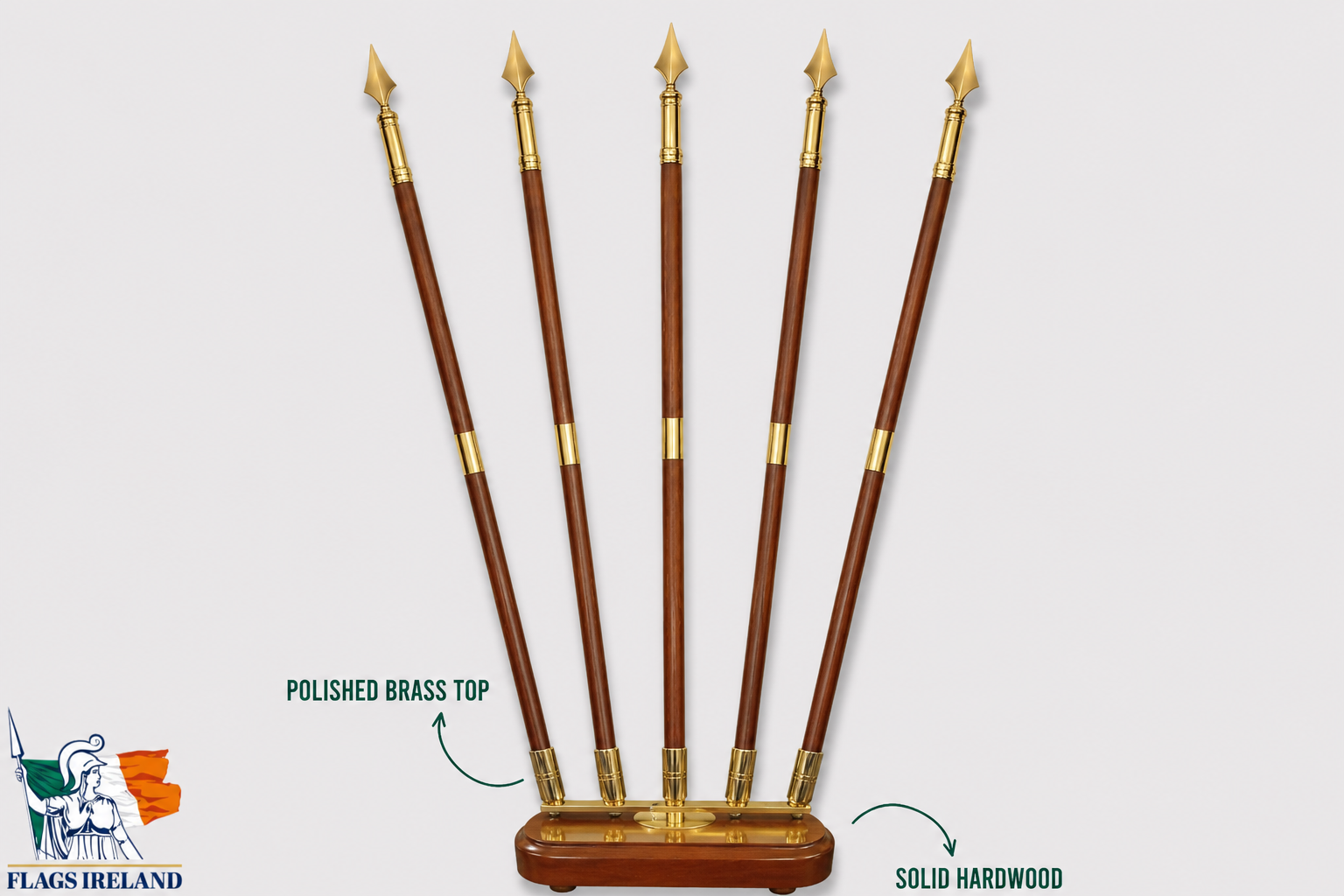 Five polished hardwood flag poles with polished brass tips arranged upright on a wooden base against a light gray background, with text callouts reading 'POLISHED BRASS TOP' and 'SOLID HARDWOOD'. A 'FLAGS IRELAND' logo appears in the lower left corner.