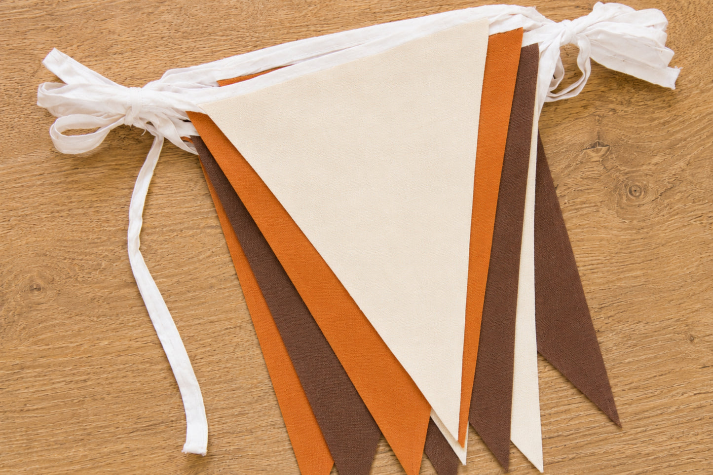 A handmade felt pennant banner with alternating cream, orange, and brown triangular flags tied to a white ribbon header, displayed on a wooden surface.