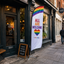Coffee Shop Storefront with a rainbow flag banner reading 'All are welcome' on a city street.