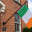 An Irish flag with green, white, and orange vertical bands hangs from a black metal wall-mounted pole attached to a red brick house exterior.