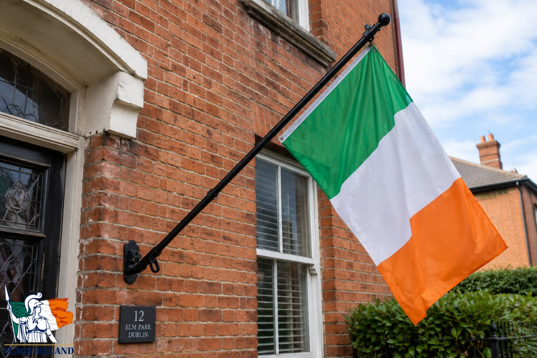 An Irish flag with green, white, and orange vertical bands hangs from a black metal wall-mounted pole attached to a red brick house exterior.