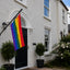 A rainbow pride flag hangs from a black metal house flagpole mounted on the exterior wall of a white stucco house. The scene includes a black front door, potted shrubs, flowering bushes, and part of the neighboring house and sky.