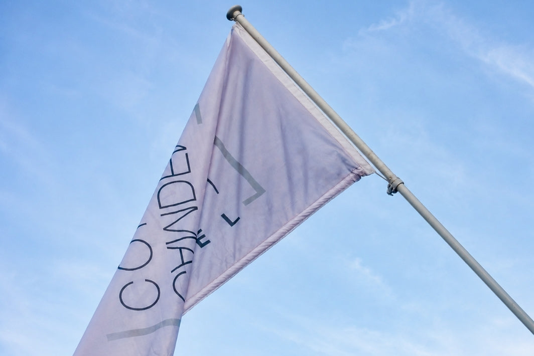 A light lavender flag with dark blue text reading 'CANDEN' and a geometric logo, attached to a silver flagpole against a bright blue sky with light clouds.