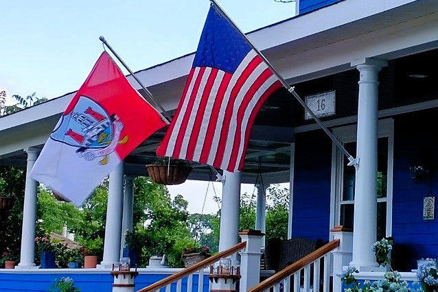 A blue house with a front porch and columns, featuring two flags hanging from the porch posts: a red, white, and blue flag on the left and the United States flag on the right. Blue hydrangea flowers are visible in pots along the porch steps.