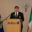 A man in a dark uniform stands behind a wooden podium with the Radisson Blu Hotel & Spa logo, flanked by two flags against a beige wall.