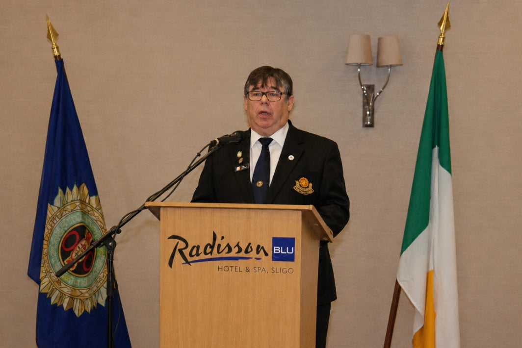 A man in a dark uniform stands behind a wooden podium with the Radisson Blu Hotel & Spa logo, flanked by two flags against a beige wall.