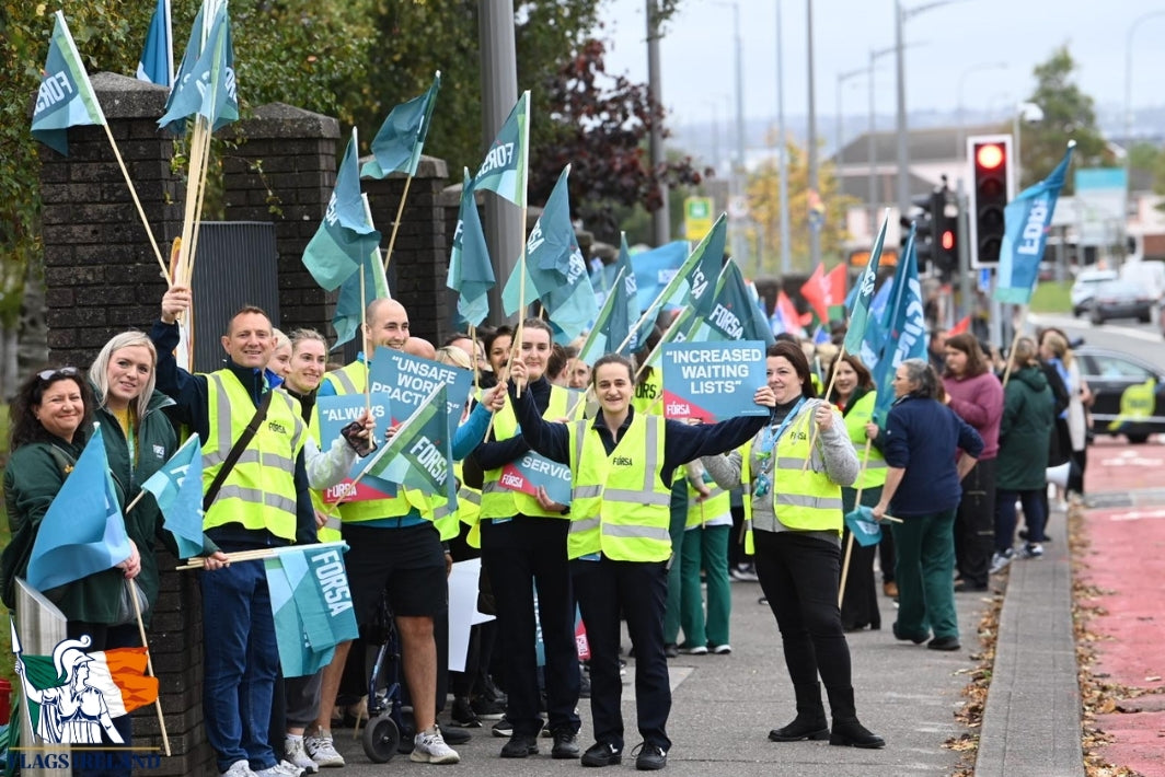 A group of healthcare workers and nurses marching outdoors on a city street, wearing high-visibility yellow vests and holding blue flags with the text 'UNFAIR PAYING WORKERS' and 'NICE ENOUGH CARE'.