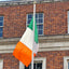 A high-quality Irish tricolour flag flying outside a historic red-brick government building. The flag is mounted on a tall flagpole, waving in the wind against a blue sky. The durable material and stitching are visible, showcasing its premium quality.