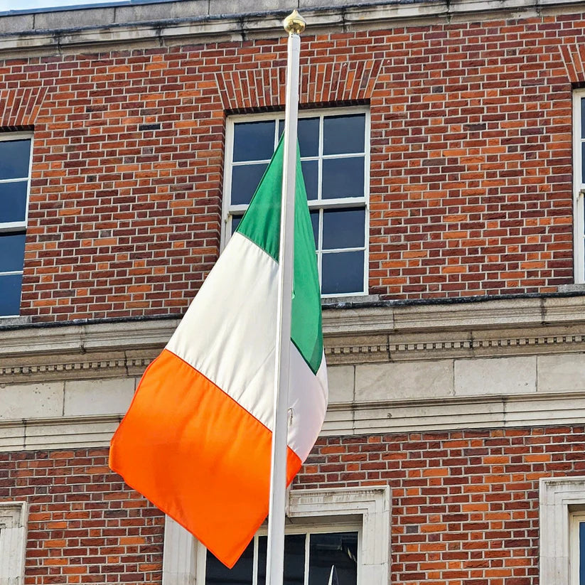 A high-quality Irish tricolour flag flying outside a historic red-brick government building. The flag is mounted on a tall flagpole, waving in the wind against a blue sky. The durable material and stitching are visible, showcasing its premium quality.