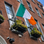 An Irish flag hangs from a metal balcony on a red brick building facade, with flower boxes and windows visible.