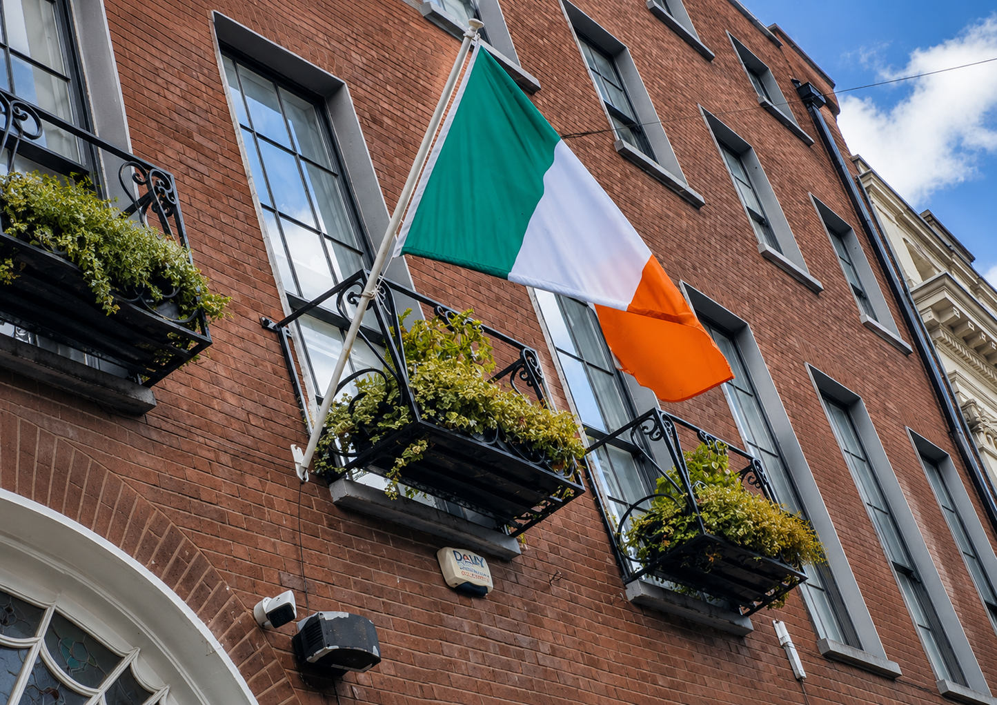 An Irish flag hangs from a metal balcony on a red brick building facade, with flower boxes and windows visible.