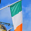 An Irish flag with green, white, and orange vertical stripes hangs from a white flagpole outside a pub. A round black Guinness sign is partially visible in the lower-left corner against a bright blue sky with light clouds.