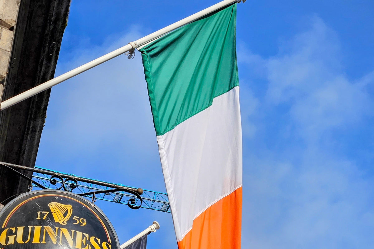 An Irish flag with green, white, and orange vertical stripes hangs from a white flagpole outside a pub. A round black Guinness sign is partially visible in the lower-left corner against a bright blue sky with light clouds.