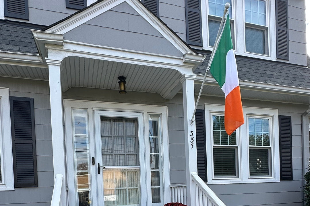 A gray house front porch decorated for autumn, with pumpkins and red flowers on the concrete steps, and an Irish flag hanging from the porch post.