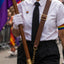 A person at a pride parade wearing a white button-up shirt, black tie, black trousers, white gloves, and a brown leather suspenders with gold hardware. They are holding a wooden pole with a rainbow pride flag and wearing a small rainbow heart pin on the chest.