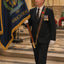 A man in a dark suit and tie stands indoors holding a blue ceremonial flag with gold fringe and the words 'Marine Corps Association.' The flag also shows an anchor emblem and gold lettering.