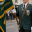 A man in a green military-style uniform holding a green flag with gold fringe and lettering stands on a street. The flag reads 'QUIS SEPARATUS ASSOCIATION.