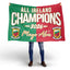 A person holding up a large rectangular banner or flag with a split green and red design, white text reading 'ALL IRELAND CHAMPIONS 2026', and the name 'Mayo Abí' in gold script. The person is only visible from the knees down, wearing blue jeans and brown shoes, against a clean white studio background.