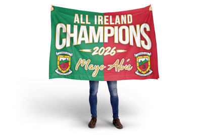 A person holding up a large rectangular banner or flag with a split green and red design, white text reading 'ALL IRELAND CHAMPIONS 2026', and the name 'Mayo Abí' in gold script. The person is only visible from the knees down, wearing blue jeans and brown shoes, against a clean white studio background.