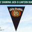 A triangular black pennant flag featuring the Oíche Shamhna mask emblem with orange and cream details, set against a light blue sky background. A dark green banner at the top reads 'OICHE SHAMHNA JACK-O-LANTERN BUNTING' and a 'Flags Ireland' logo is visible in the bottom left corner.