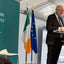 A man in a dark suit stands at a green podium speaking into a microphone, with flags including the Irish flag and EU flag beside a teal sign reading 'Socialistáis na hEireann Government of Ireland'.