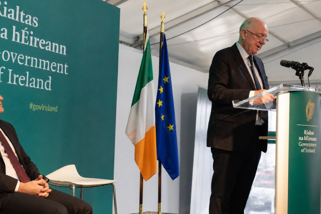 A man in a dark suit stands at a green podium speaking into a microphone, with flags including the Irish flag and EU flag beside a teal sign reading 'Socialistáis na hEireann Government of Ireland'.