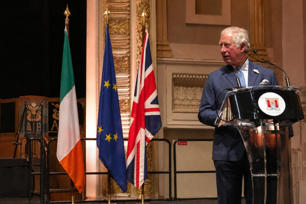 Prince Charles speaking at a podium with Irish, EU, and UK flags in the background.