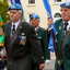 A group of elderly men in military-style uniforms marching outdoors. The visible uniforms are dark green with blue berets, medals, and shoulder straps, and several carry flags.