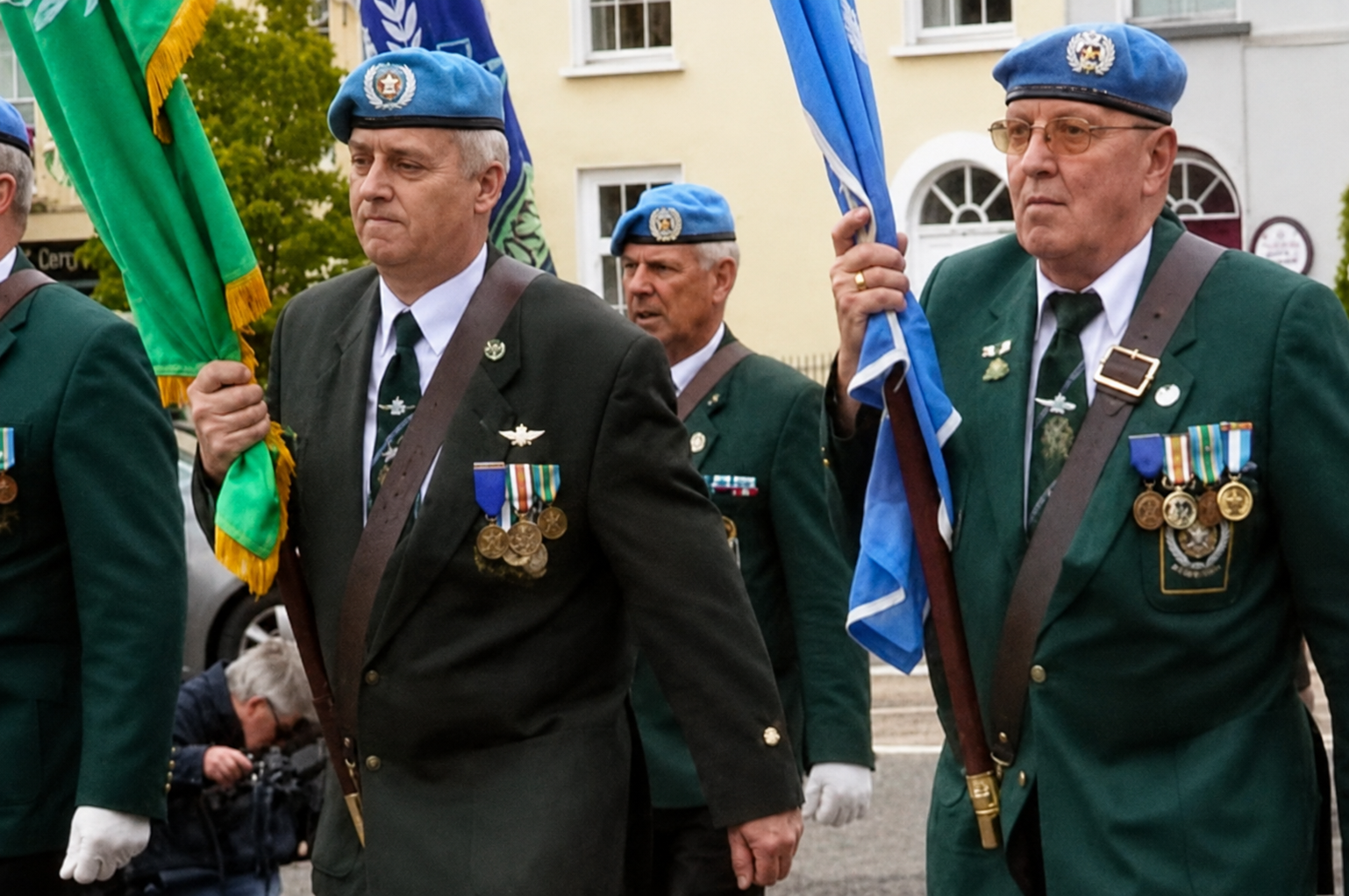 A group of elderly men in military-style uniforms marching outdoors. The visible uniforms are dark green with blue berets, medals, and shoulder straps, and several carry flags.