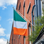 An Irish flag with green, white, and orange vertical bands hangs from a metal pole mounted on a red brick building under a blue sky with clouds.