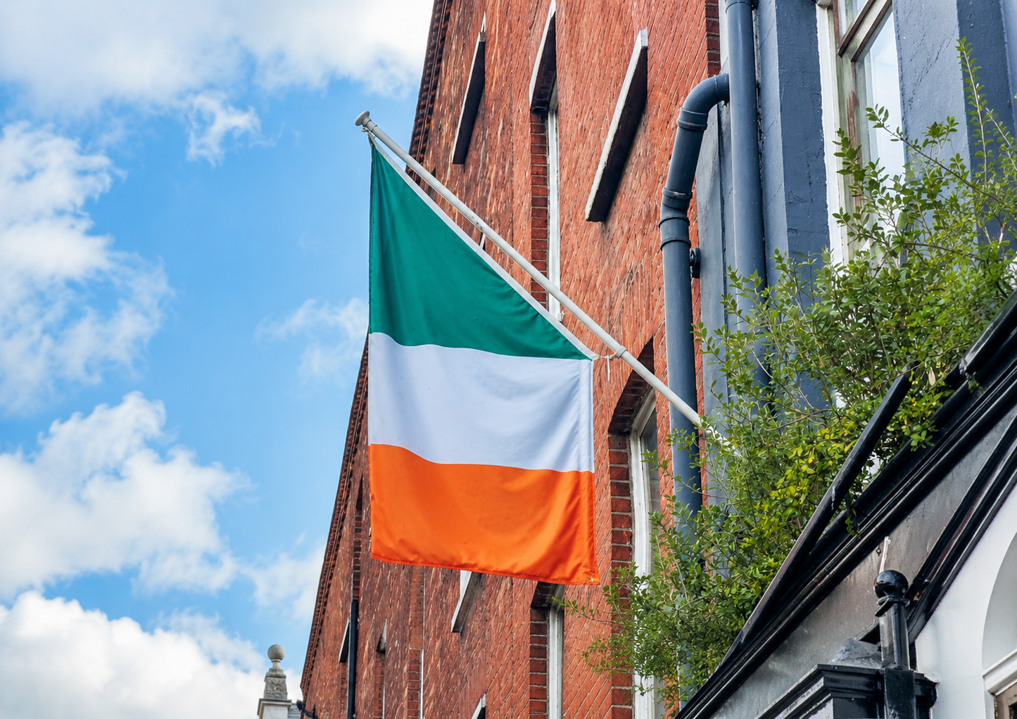 An Irish flag with green, white, and orange vertical bands hangs from a metal pole mounted on a red brick building under a blue sky with clouds.
