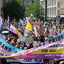 A crowded outdoor street protest or pride march with many people holding signs and banners. Prominent visible text includes 'TRANS & LGB+ PRIDE', 'NO PRIDE FOR CENSURE', 'DELIBERATION FOR ALL!', and 'HEALTHCARE NOW'.