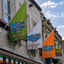 Three decorative flags for 'The Boomerang Bar & Bistro' celebrating 2016, in green, white, and orange, hanging from a building facade under a blue sky with clouds.