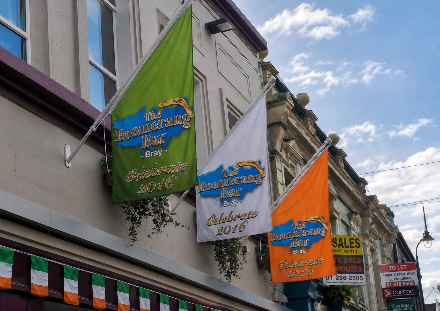 Three decorative flags for 'The Boomerang Bar & Bistro' celebrating 2016, in green, white, and orange, hanging from a building facade under a blue sky with clouds.