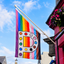 A pride flag featuring a large white letter 'P' decorated with paint palette dots and vertical rainbow stripes, hanging from a building with pink walls and flower boxes under a blue sky.