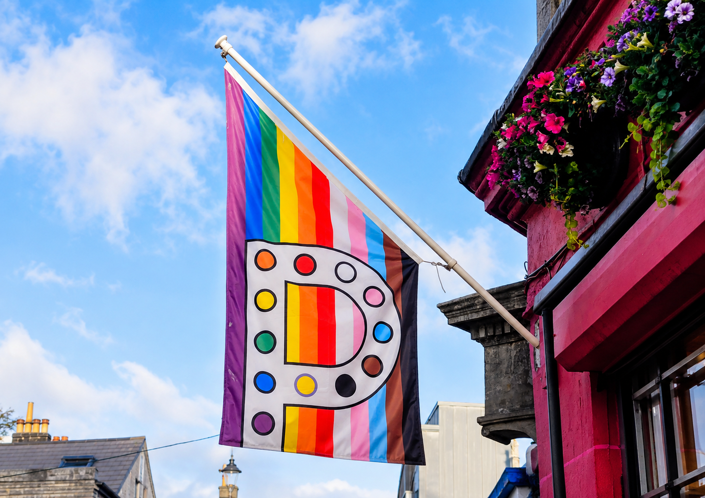 A pride flag featuring a large white letter 'P' decorated with paint palette dots and vertical rainbow stripes, hanging from a building with pink walls and flower boxes under a blue sky.