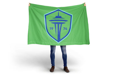 Person holding a green flag with a blue logo featuring the Space Needle and the year 1974 on a white .background. Seattle Sounders FC Flag- New 2024 Crest