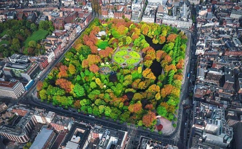 Aerial view of a large park, Stephens green in Dublin with dense green, yellow, orange, and red trees, surrounded by city streets and buildings.