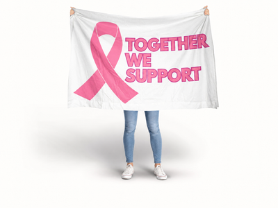 A person standing against a light gray studio background holds up a large white banner or flag with a pink awareness ribbon and the words “TOGETHER WE SUPPORT” in pink. The person’s head is cropped out, but their legs and shoes are visible below the banner. Breast Cancer Flag Banner