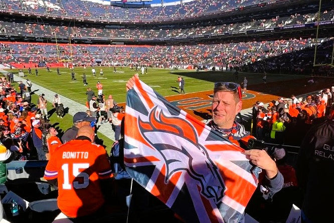 A man in a stadium stands holding a large Denver Broncos flag, with a football field and packed arena visible in the background.