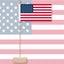 A small table flag of American on a thin pole with a wooden base, shown in front of a larger American flag background.