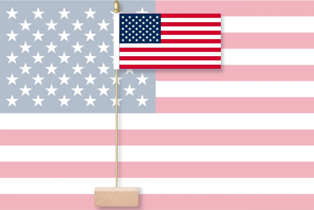 A small table flag of American on a thin pole with a wooden base, shown in front of a larger American flag background.