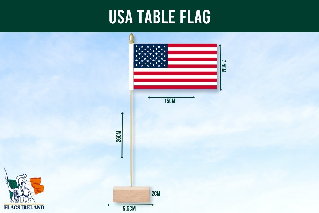 USA table flag with a wooden base, metal pole, and gold finial, shown with dimension labels against a light blue sky background. A dark green banner at the top reads 'USA TABLE FLAG,' and a 'Flags Ireland' logo appears in the lower left.