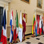 A row of international flags displayed on poles in an elegant indoor hall with cream walls, framed artwork, and a black-and-white checkered floor.